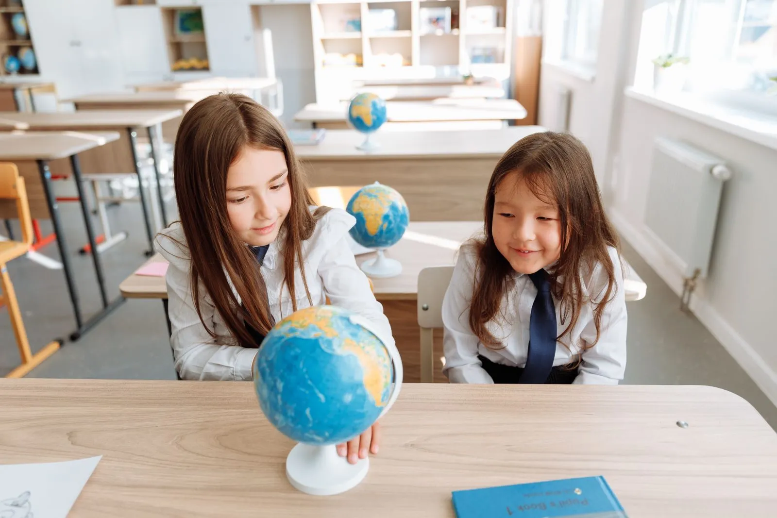 Two girls studying geography in a bright classroom with a globe on the table.